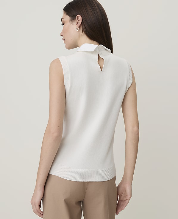 A woman in a white sleeveless top and tan pants stands against a plain, light-colored background.