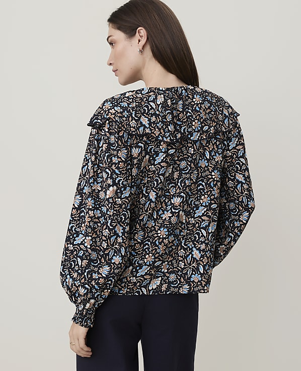 A woman in a dark floral blouse with a ruffled collar stands against a plain, light-colored background.