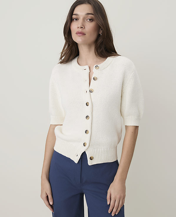 A woman in a cream-colored cardigan and blue pants stands against a plain, light gray background.