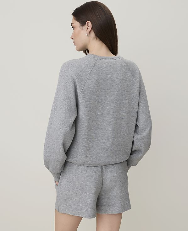 A woman in a grey sweatshirt and shorts stands with her back to the camera against a plain grey background.