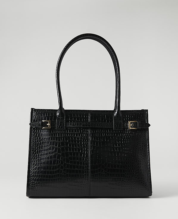 A black crocodile-embossed tote bag with gold buckle accents sits against a plain, light gray background.