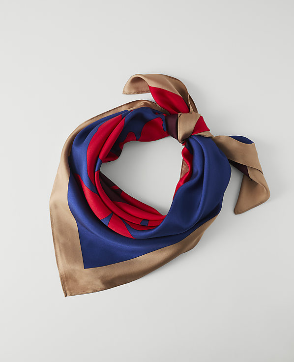 A silk scarf with a red and blue pattern and tan trim is tied in a knot against a plain white background.