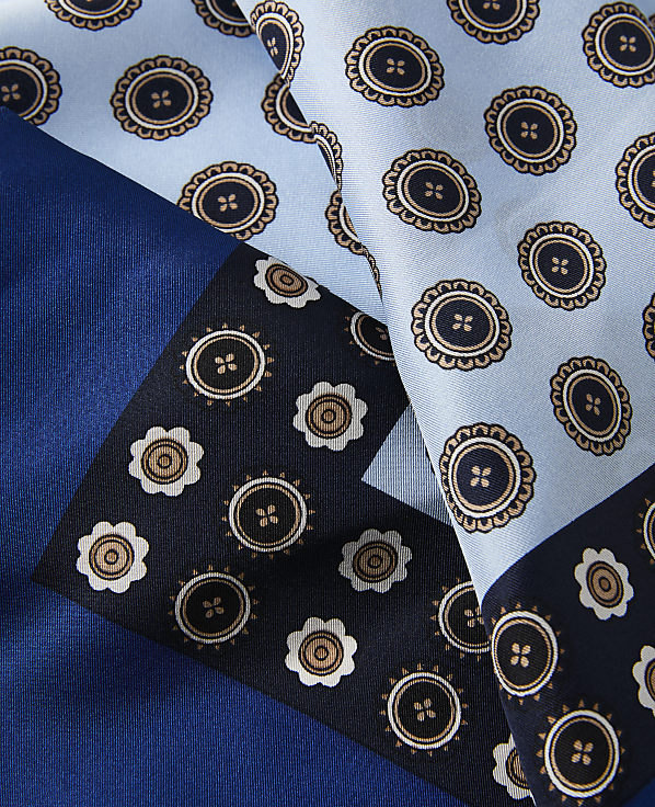 A close-up of a silk scarf with a geometric pattern of circular motifs in shades of blue and brown is laid out against a solid blue background.