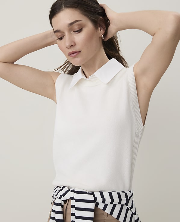 A woman with her hands behind her head wears a white sleeveless collared top and a striped sweater tied around her waist against a plain, light background.