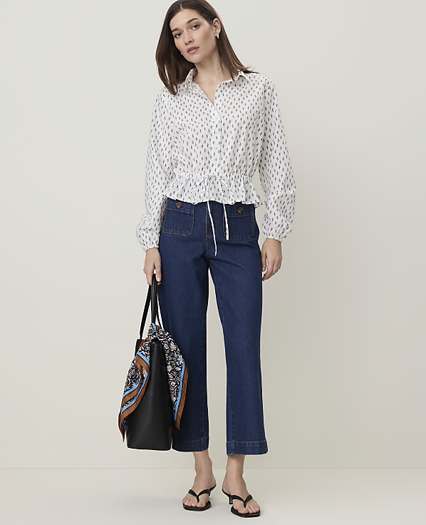 A woman in a patterned blouse and dark jeans stands with a tote bag in front of a plain white background.
