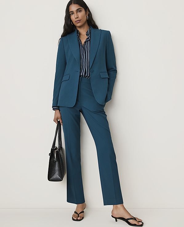 A woman in a teal pantsuit and black heels stands holding a black handbag against a plain white background.