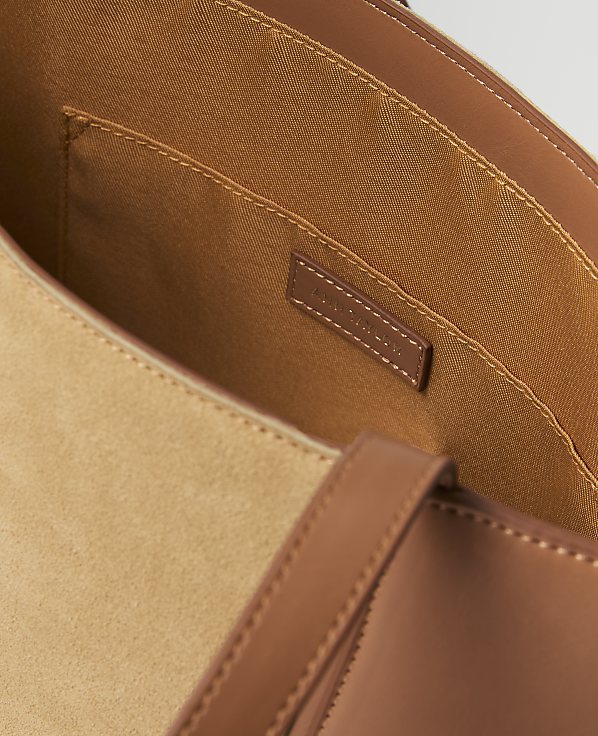 A close-up view of a tan suede tote bag's interior with brown leather trim and straps in the foreground, and a small brand tag visible in the background.
