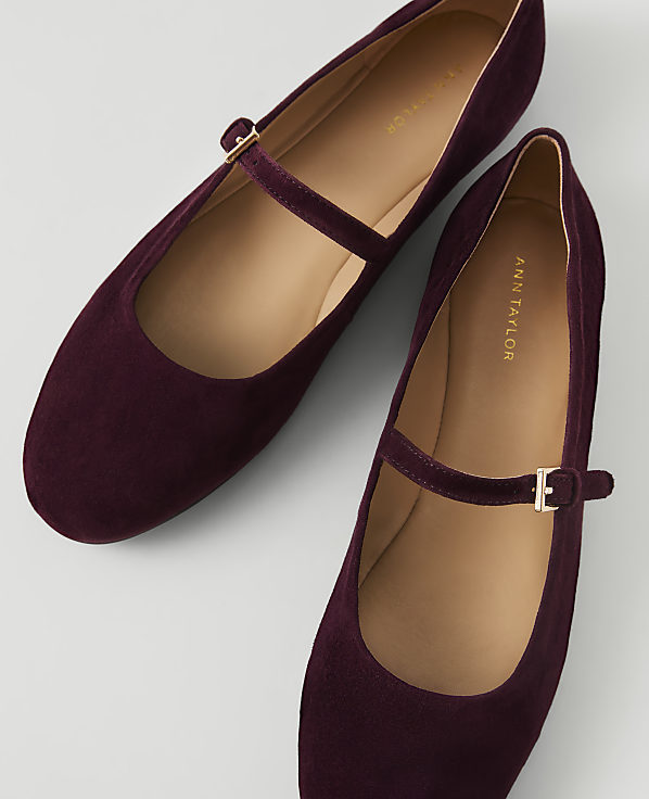 A pair of deep burgundy suede Mary Jane flats with gold buckles rests on a light gray background.