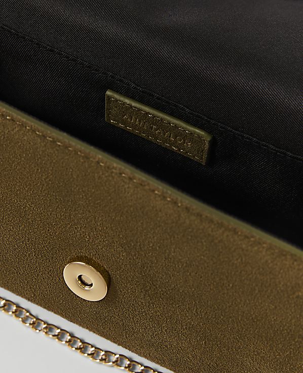 A close-up of an olive green suede clutch with a gold chain strap in the foreground, and a black interior with a brand tag in the background.