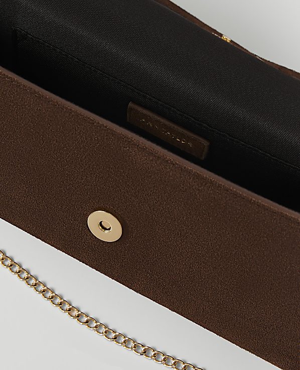 A brown clutch with a gold chain strap in the foreground is open to reveal a black interior with a brand tag in the background.