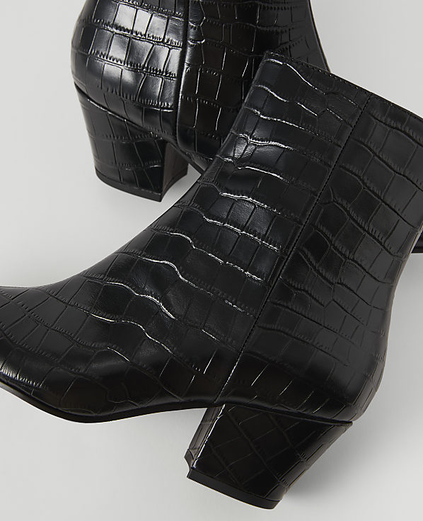 Black, croc-embossed ankle boots with block heels are arranged on a white surface.