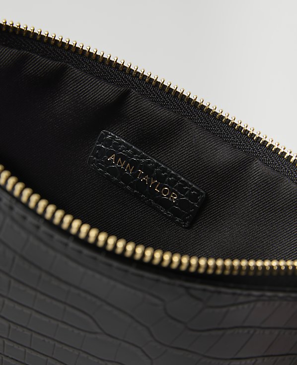 A black, crocodile-embossed clutch with a gold zipper is open to reveal a black interior with an "ANN TAYLOR" logo.