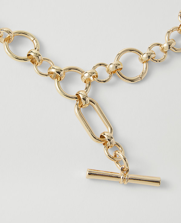 A gold toggle clasp necklace with circular links in the foreground rests on a plain white background.