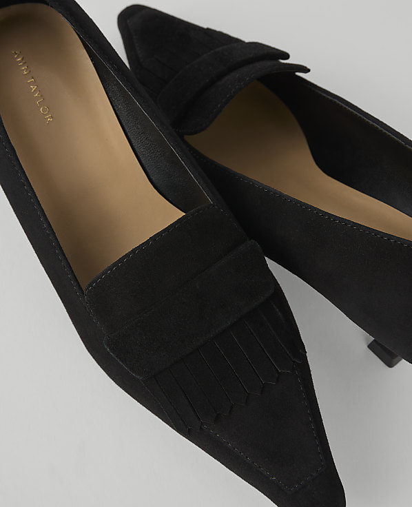 A pair of black suede pointed-toe loafers with fringe detail are in the foreground, against a plain white background.