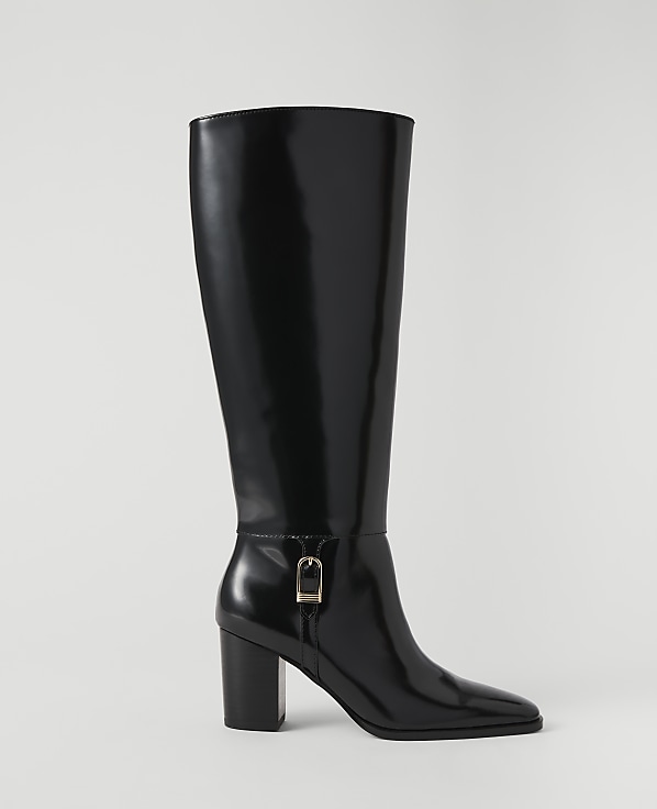 A shiny black knee-high boot with a gold buckle stands against a plain white background.