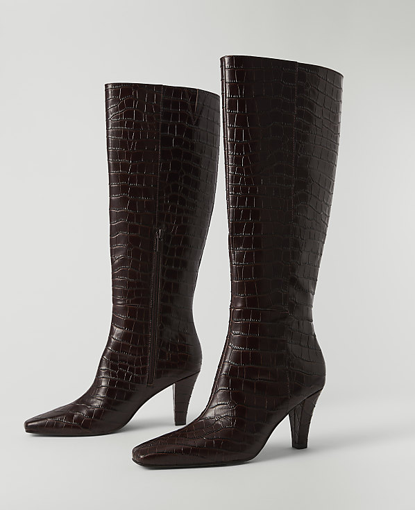 A pair of dark brown, croc-embossed leather knee-high boots with pointed toes and stiletto heels stand on a plain white surface against a white background.
