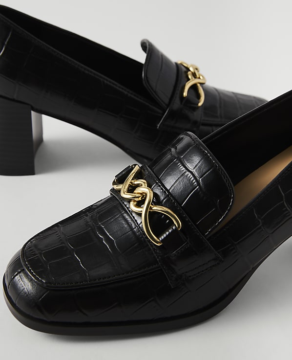Black croc-embossed heeled loafers with gold chain accents are in the foreground, with another shoe slightly out of focus in the background.