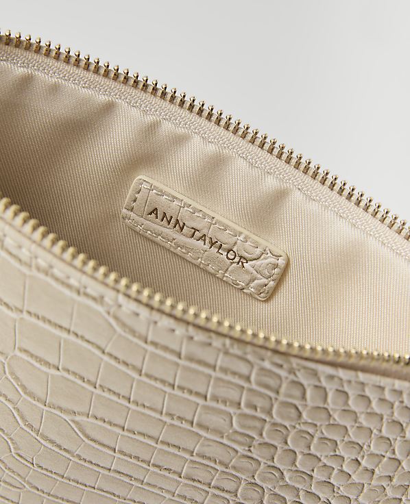 A close-up of a cream-colored, croc-embossed clutch bag with a gold zipper is shown against a plain white background.