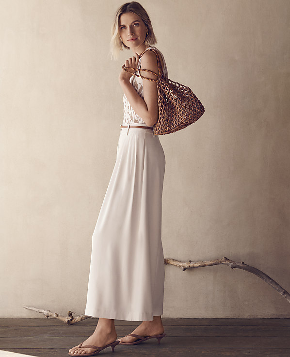 A woman in a white top and flowing white pants stands with a woven bag, against a textured wall with a branch.