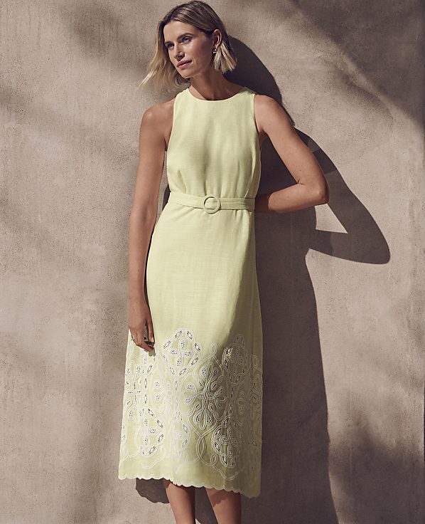A woman in a pale yellow sleeveless dress with an embroidered hem stands against a textured, sunlit wall.