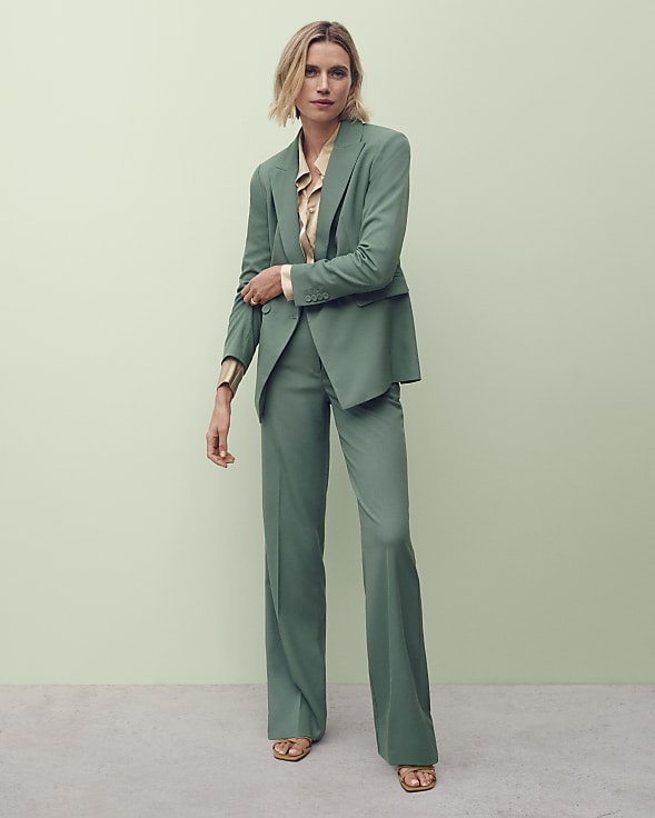 A woman in a green pantsuit stands against a pale green background.