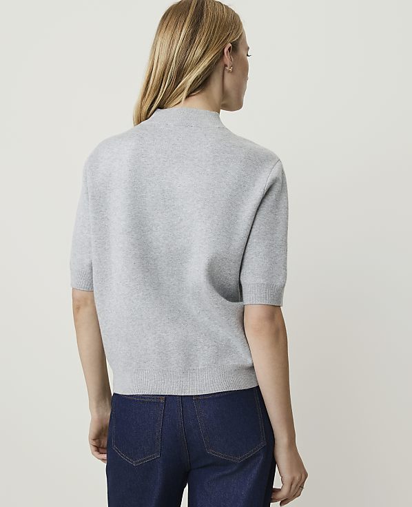 A woman in a grey short-sleeved sweater and dark jeans stands against a plain, light-colored background.