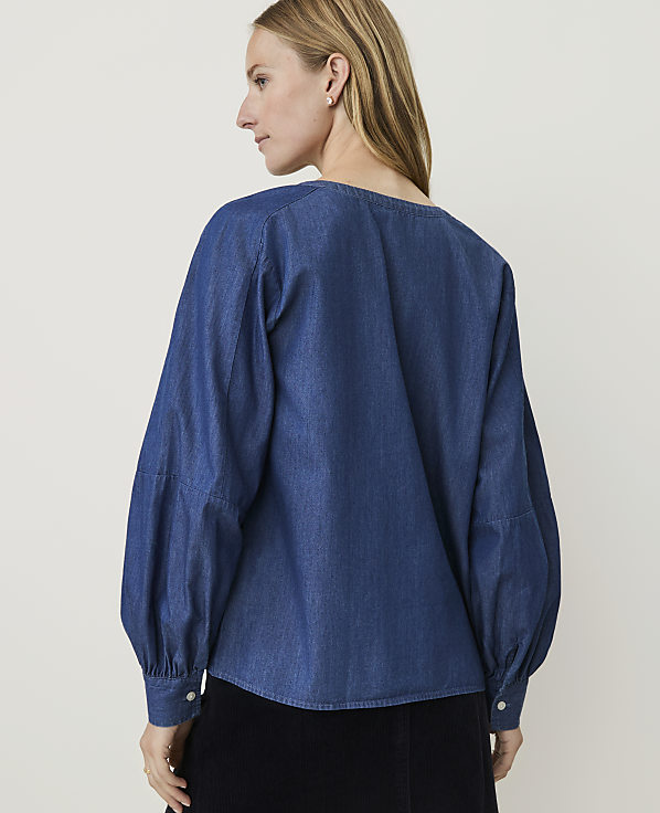 A woman in a blue denim blouse with voluminous sleeves stands against a plain white background.
