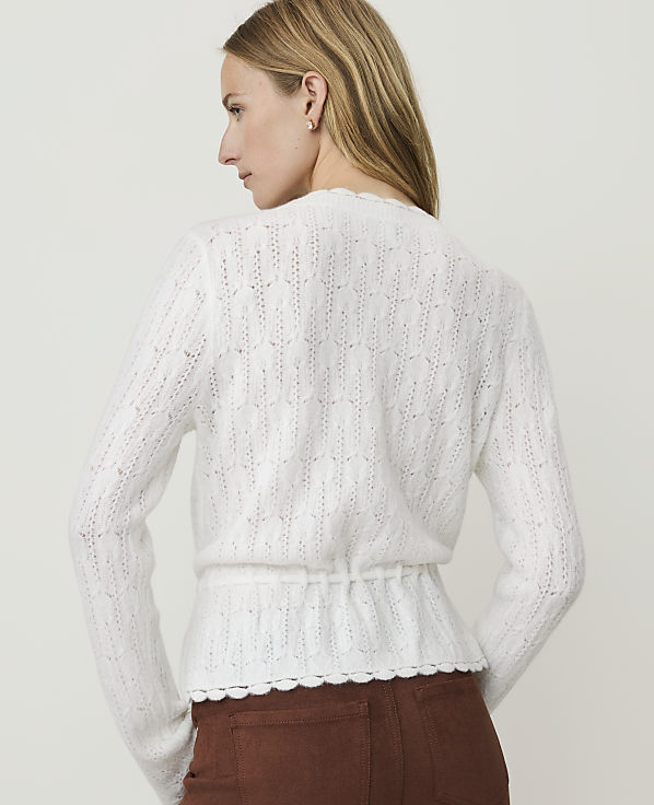 A woman in a white, textured cardigan with a cinched waist stands against a plain, light background.