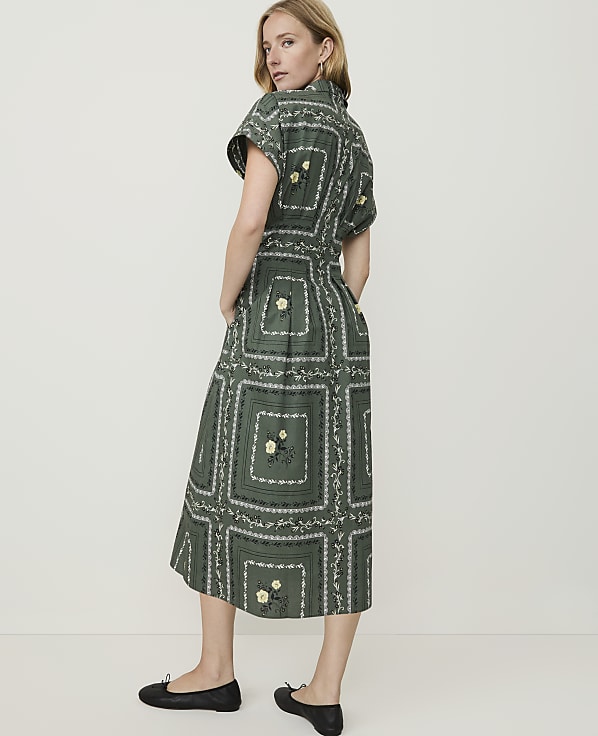 A woman in a green, patterned midi dress and black flats stands against a plain, light-colored background.