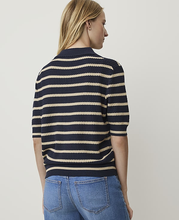A woman in a striped polo shirt and jeans stands against a plain white background.
