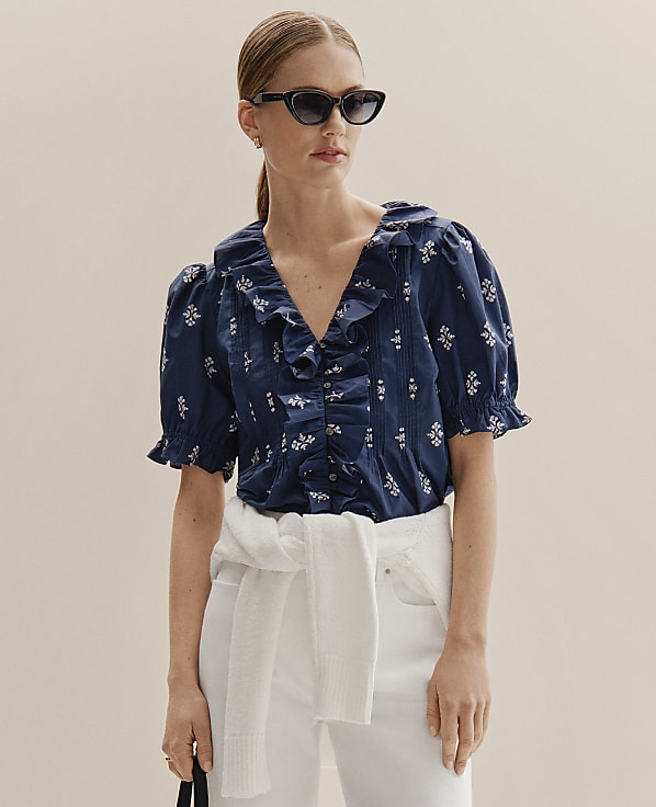 A woman in a navy floral blouse and white pants stands against a plain, light-colored background.