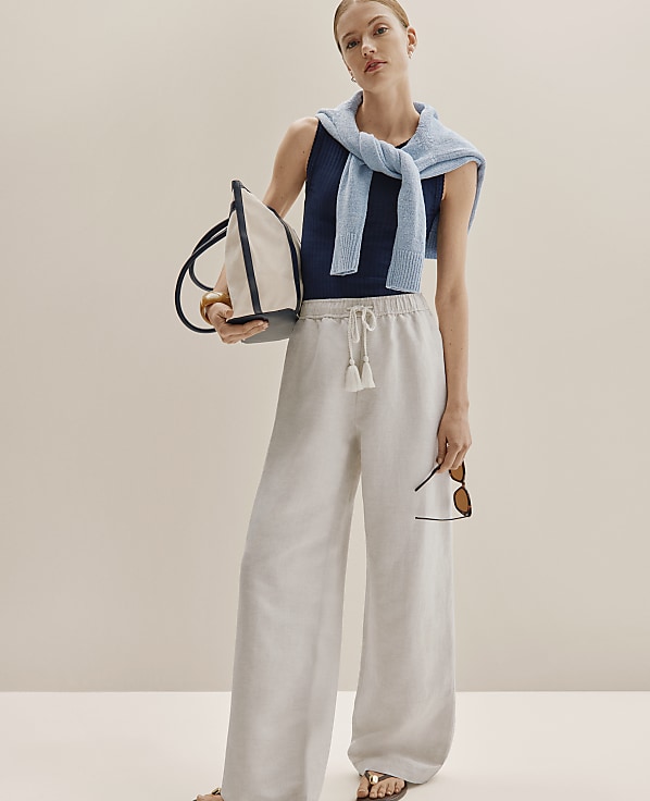 A woman in wide-leg linen pants and a striped tank top stands against a plain, light-colored background, holding a tote bag and sunglasses.
