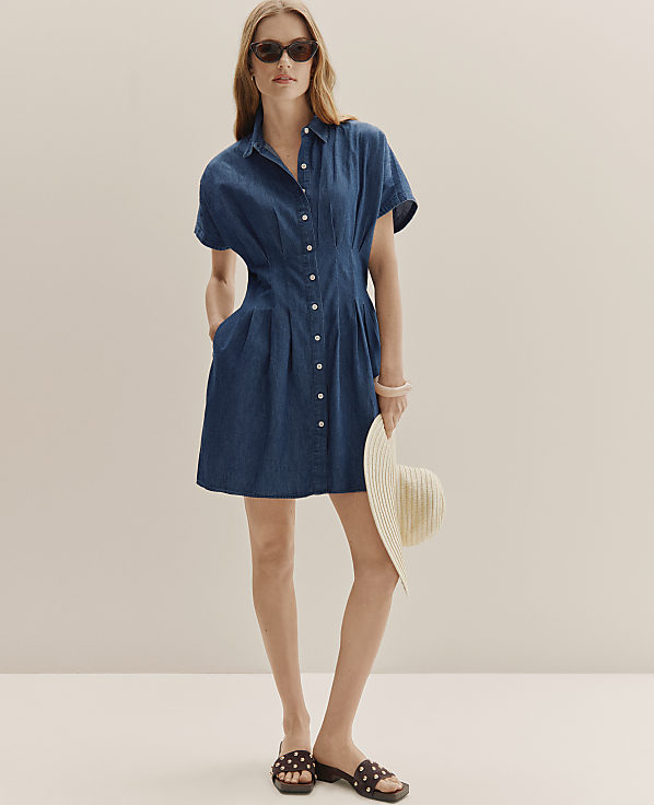 A woman in a denim dress and sunglasses stands with a hat in her hand against a plain, light-colored background.