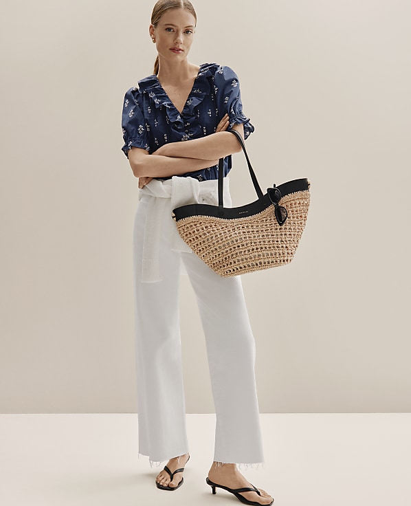 A woman in white pants and a floral top stands with a straw bag in front of a plain background.