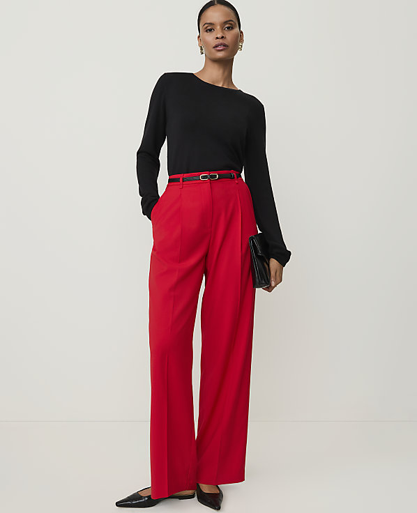 A woman in a black sweater and red pants stands against a plain white background, holding a black clutch.