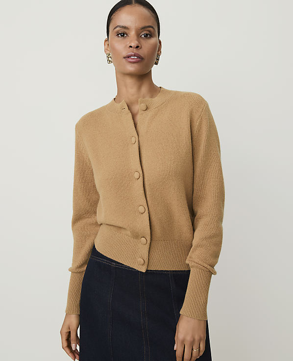 A woman in a tan cardigan and dark skirt stands against a plain background.