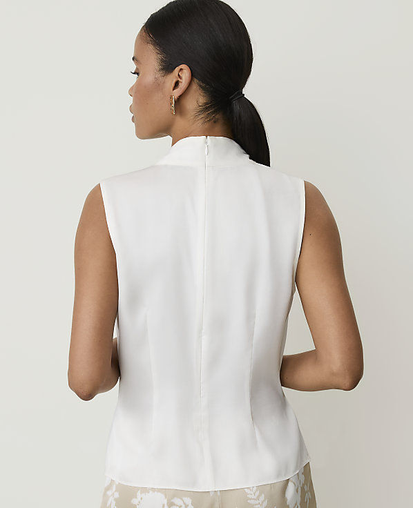 A woman in a white sleeveless top stands against a plain, light background.