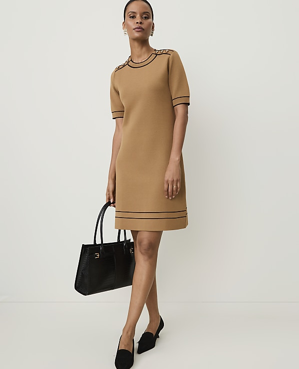 A woman in a tan knit dress with black trim stands holding a black handbag against a plain white background.