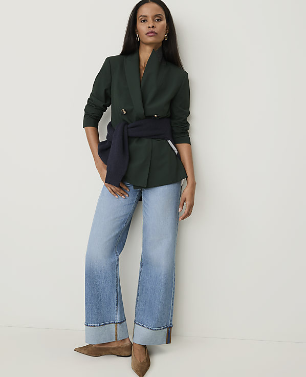 A woman in a dark green blazer and light wash jeans stands against a plain white background.