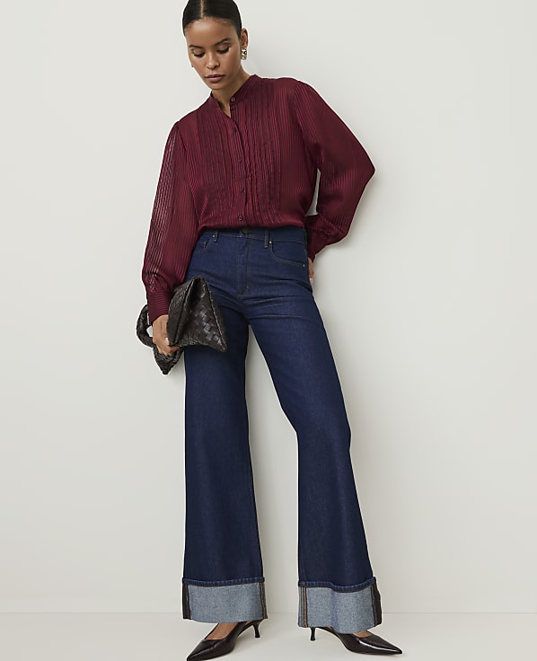 A woman in a maroon blouse and dark flared jeans stands against a plain white background, holding a dark clutch.