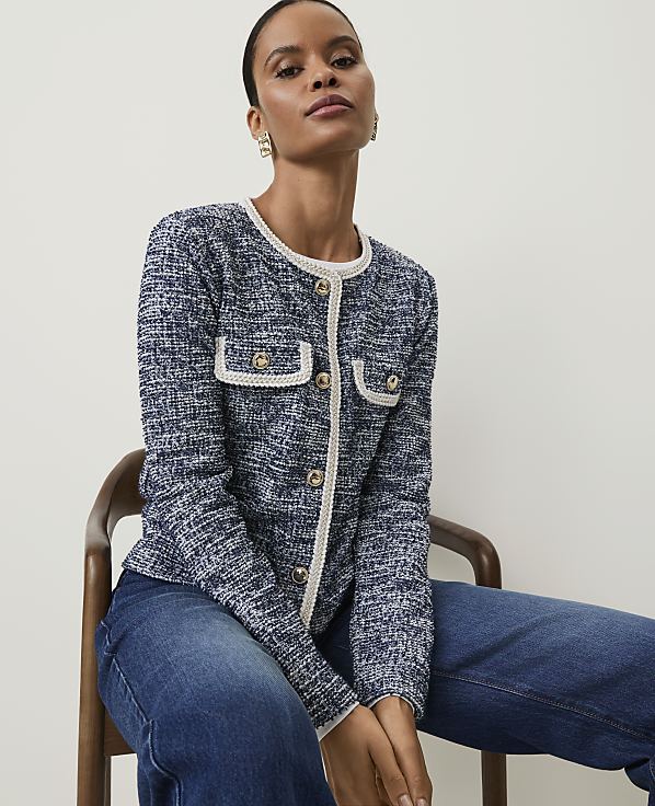 A woman in a blue tweed jacket and jeans sits on a wooden chair against a plain white background.