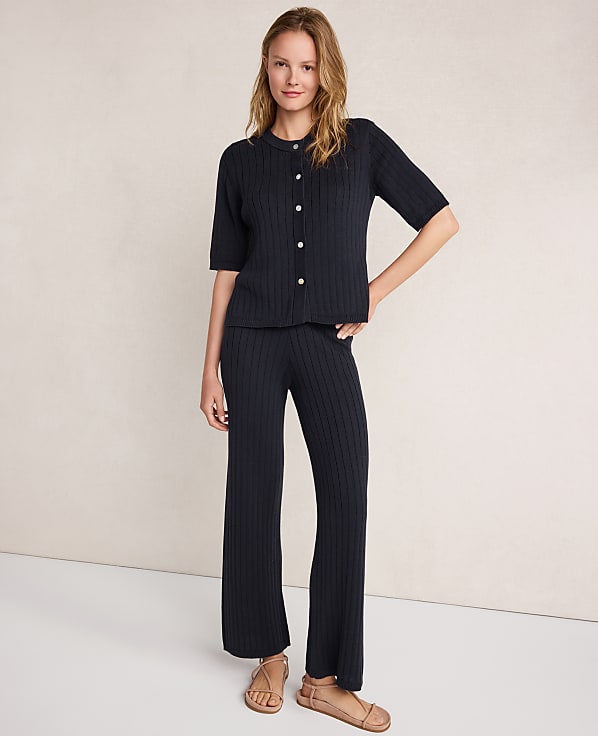 A woman in a dark ribbed knit cardigan and matching pants stands against a plain, light-colored background.