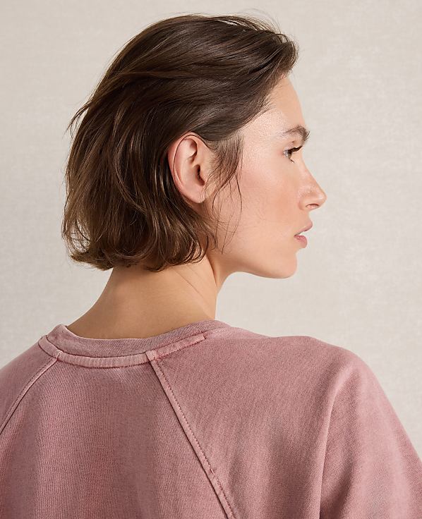 A woman with short brown hair in a pink sweatshirt is shown against a plain, light-colored background.