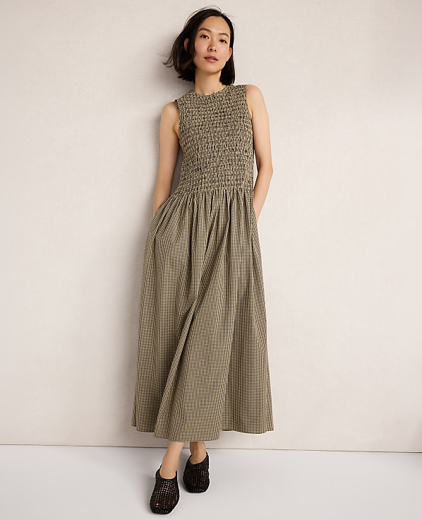 A woman in a smocked olive green dress stands against a plain, light-colored wall.
