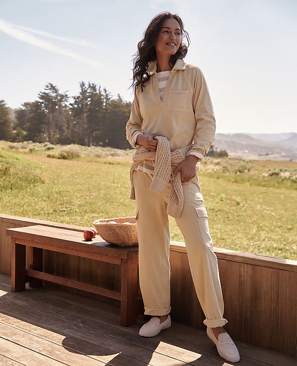 A woman in a beige jumpsuit stands on a wooden deck in front of a basket of apples, with rolling hills and trees in the background under a bright sky.