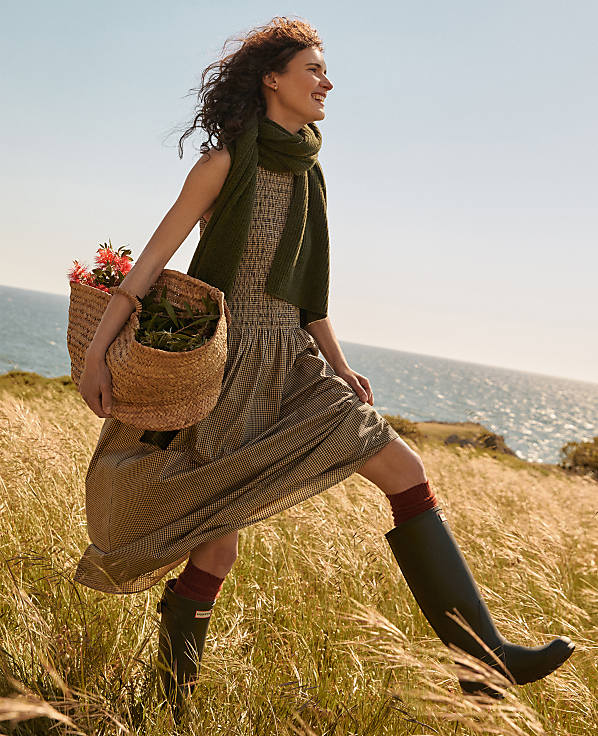 A woman in a dress and boots walks through tall grass carrying a basket, with the ocean and sky in the background.