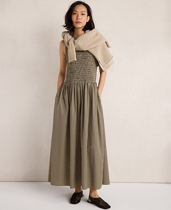 A woman in a green smocked dress and a beige sweater draped over her shoulders stands against a plain, light-colored background.