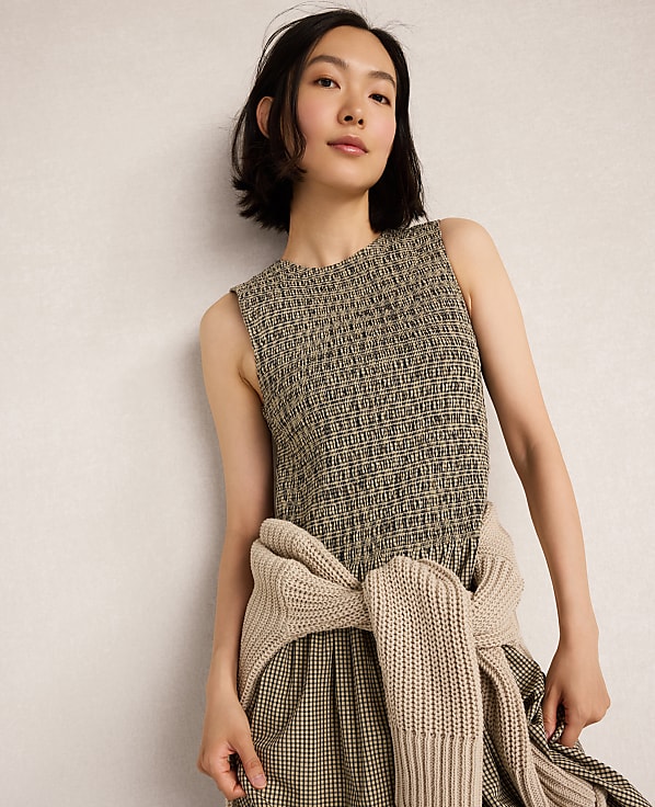 A woman in a textured sleeveless dress and a beige sweater tied around her waist stands against a plain, light-colored wall.