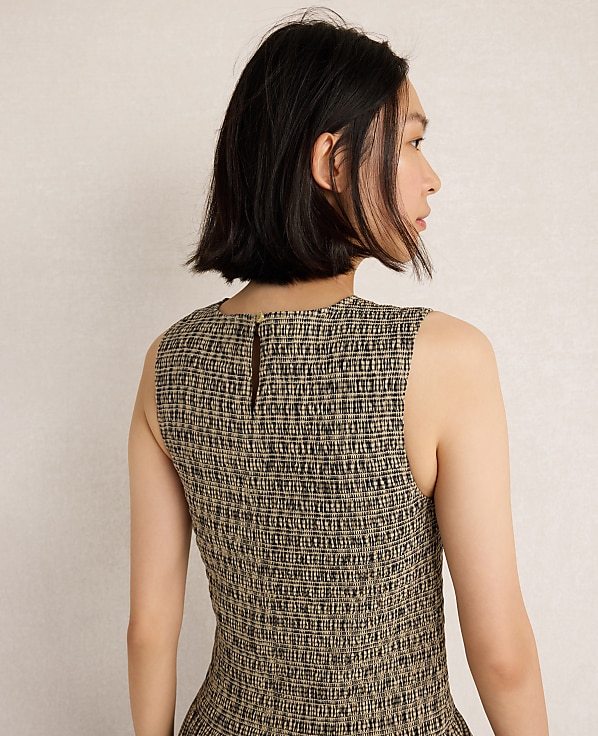 A woman with short dark hair, seen from the back, wears a textured sleeveless dress against a plain, light-colored background.
