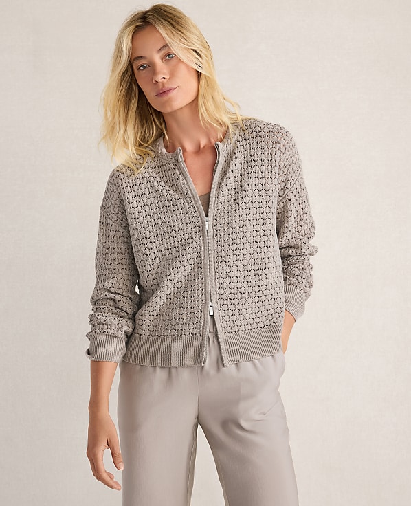 A blonde woman in a textured gray zip-up cardigan and light gray pants stands against a neutral, textured background.